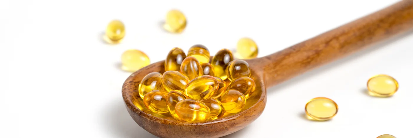 A wooden spoon heaped with golden omega-3 softgel capsules, with more capsules scattered on a white surface.